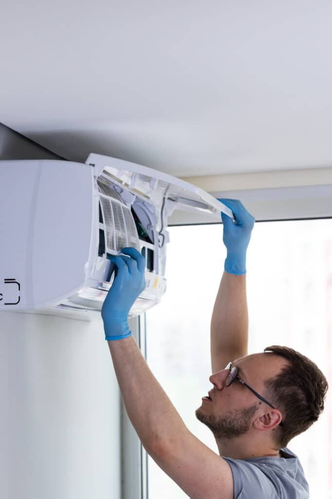 Entretien climatiseur mural : nettoyage et maintenance CVC pro. Homme en lunettes et gants bleus inspectant un climatiseur mural ouvert, révélant les filtres. Entretien et maintenance CVC.