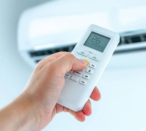 Main tenant une télécommande de climatiseur, réglant la température à 26°C. L'unité de climatisation murale est visible en arrière-plan.