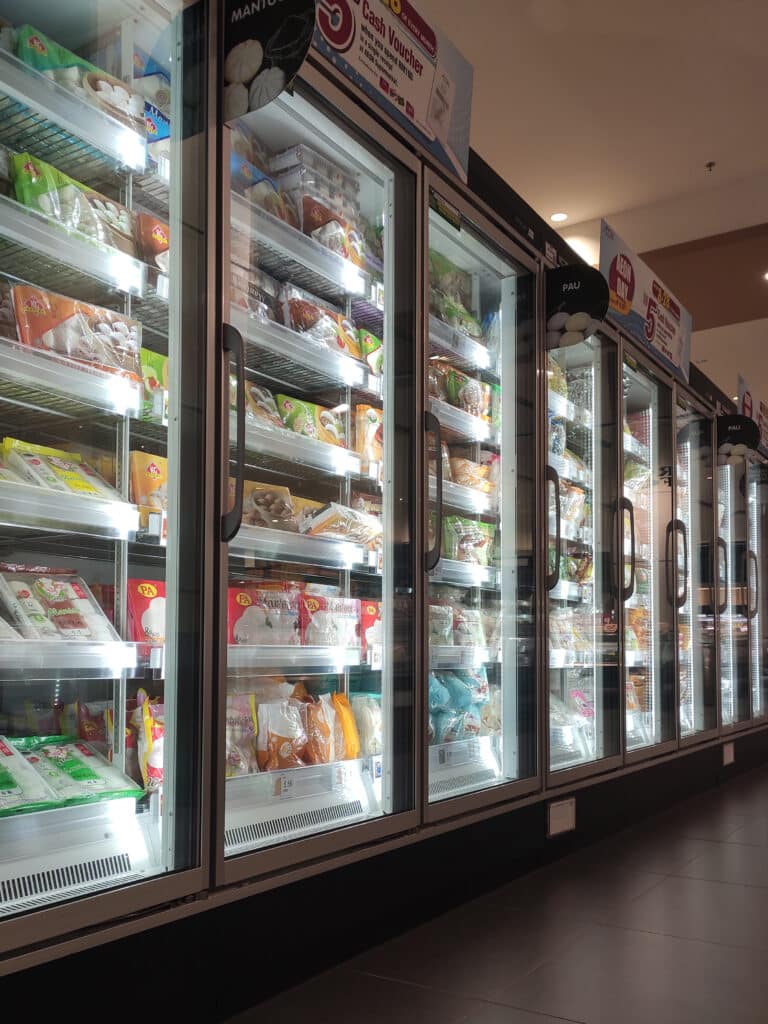 Supermarché AEON: Congélateurs de Mantou et Pâtisseries Surgelées Allée de congélateurs vitrés lumineux d'un supermarché AEON, présentant Mantou et pâtisseries surgelées. Un panneau mentionne un 'Cash Voucher' pour 100 RM.